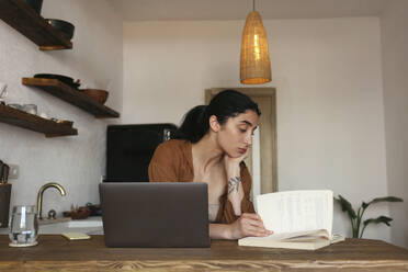 Young woman studying with book and laptop on table at home - SYEF00399