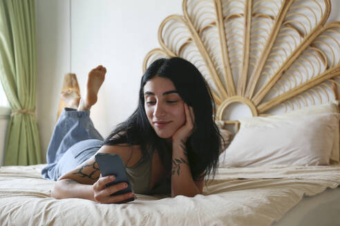 Junge Frau mit Smartphone auf dem Bett liegend zu Hause - SYEF00412