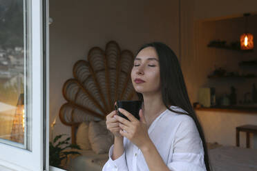 Junge Frau mit geschlossenen Augen hält Kaffeetasse am Fenster - SYEF00420