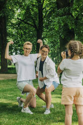 Daughter photographing playful lesbian couple posing with flexing muscles at park - VSNF01030