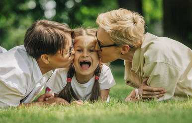 Lesbisches Paar küsst Tochter im Gras liegend im Park - VSNF01040