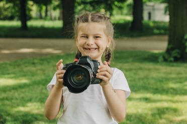 Cheerful girl holding camera at park - VSNF01046