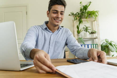 Smiling young businessman looking at document on desk in office - OSF01600