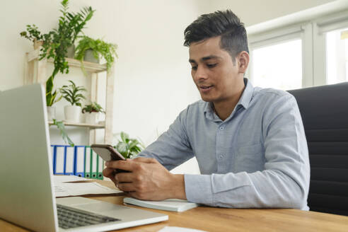 Geschäftsmann sitzt mit Laptop und benutzt ein Smartphone am Schreibtisch im Büro - OSF01617