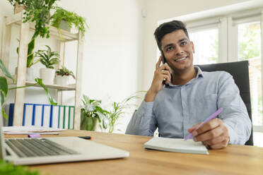 Smiling young businessman talking on smart phone and writing notes in office - OSF01619