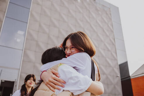 Glückliches Mädchen, das seine Freundin umarmt, vor einem Schulgebäude stehend - MDOF01258