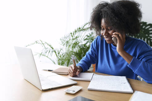 Afro freelancer talking through smart phone and writing on note pad with laptop at desk - AAZF00696