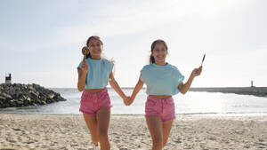 Zwillingsschwestern halten Hände mit Lutscher Süßigkeiten und laufen am Strand - ASGF03655