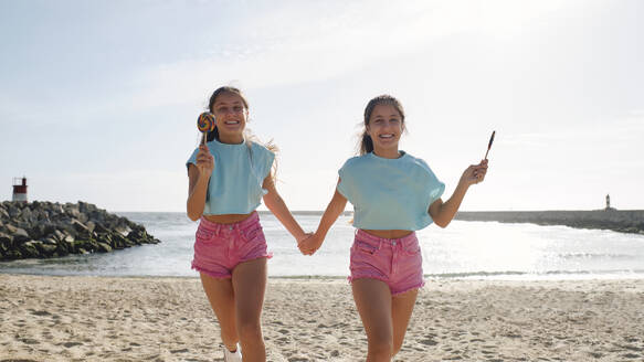 Twin sisters holding hands with lollipop candy and running at beach - ASGF03655