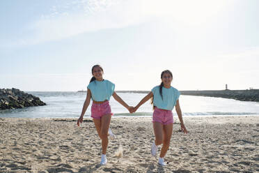 Sorglose Zwillingsschwestern, die sich an den Händen halten und am Strand spazieren gehen - ASGF03663
