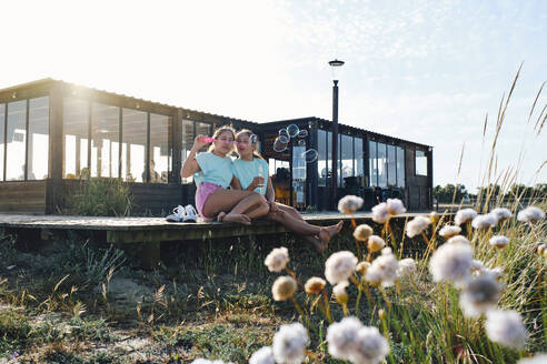 Twin sisters blowing bubbles sitting on pier at beach - ASGF03665