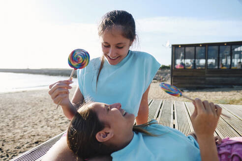 Glückliche Zwillingsschwestern genießen Lollipop-Bonbons auf dem Steg am Strand - ASGF03671