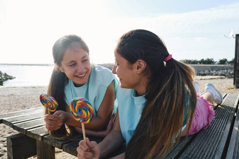 Lächelnde Zwillingsschwestern liegen auf dem Steg und halten Lutschbonbons am Strand - ASGF03675