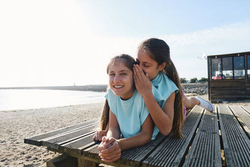 Mädchen flüstert seiner Zwillingsschwester zu, die auf einem Steg am Strand liegt - ASGF03676