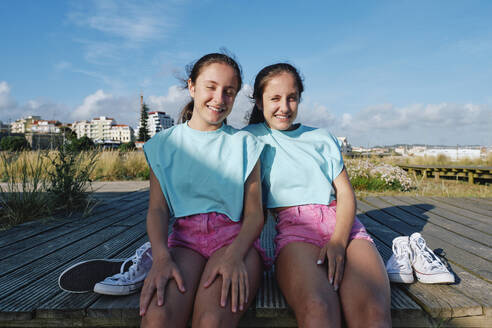 Twin sisters wearing matching outfits sitting on jetty at beach - ASGF03680