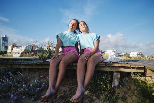 Twins sisters wearing matching outfits relaxing on jetty at beach - ASGF03681