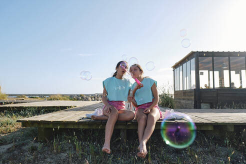 Twin sisters blowing bubbles sitting on jetty at beach - ASGF03682