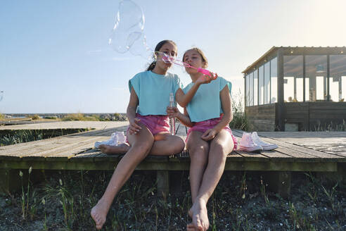 Twin sisters blowing bubbles on jetty at beach - ASGF03683