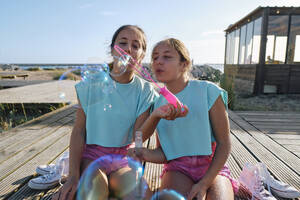 Zwillingsschwestern blasen am Strand sitzend Seifenblasen - ASGF03684