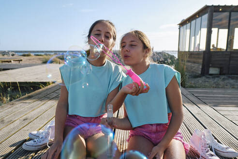 Twin sisters blowing bubbles sitting at beach - ASGF03684