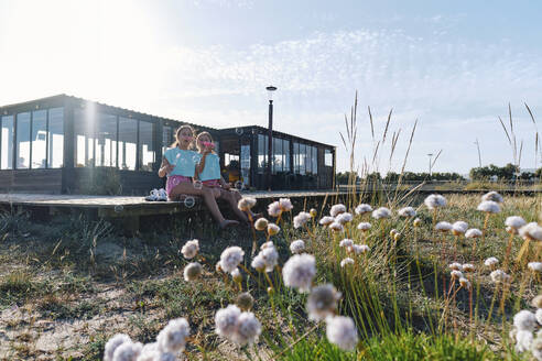 Twin sisters blowing bubbles sitting on jetty by log cabin at beach - ASGF03685