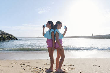 Zwillingsschwestern in passenden Outfits stehen Rücken an Rücken mit Spritzpistolen am Strand - ASGF03690