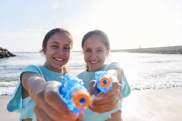 Smiling twin sisters holding squirt guns at beach - ASGF03693