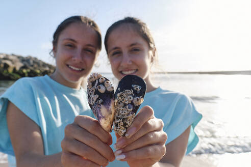 Smiling twin sisters holding seashells at beach - ASGF03699