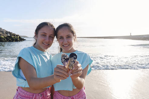 Smiling twin sisters holding seashells near sea at beach - ASGF03700