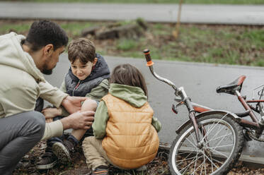Vater untersucht die Wunde seines Sohnes am Straßenrand sitzend neben dem Fahrrad - ANAF01525