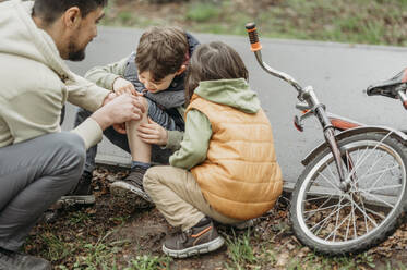 Vater, der die Wunde seines Sohnes tröstet, sitzt am Straßenrand in der Nähe des Fahrrads - ANAF01526