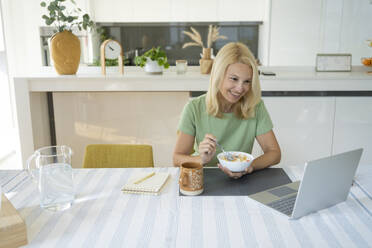 Smiling freelancer holding breakfast bowl in front of laptop at home - SVKF01419