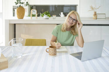 Smiling freelancer wearing eyeglasses in front of laptop at home - SVKF01421