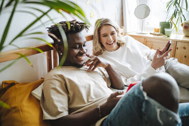 Smiling woman looking at boyfriend's smart phone relaxing on bed - MDOF01317