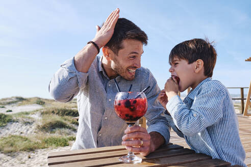 Fröhlicher Vater hält ein Glas Saft in der Hand und sein Sohn isst am Tisch - ASGF03736