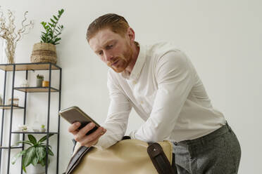 Businessman using smart phone and leaning on bag in office - OSF01646