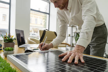 Geschäftsmann, der auf einem Notizblock schreibt und ein Solarpanel am Schreibtisch untersucht - OSF01648