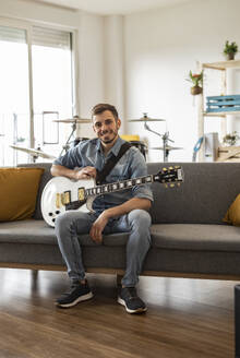 Glücklicher junger Gitarrist sitzt mit Gitarre auf dem Sofa zu Hause - JCCMF10481