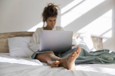 Woman using laptop sitting on bed at home - IKF00819