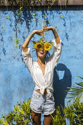 Smiling woman hiding face with sunflowers in front of wall - IKF00833