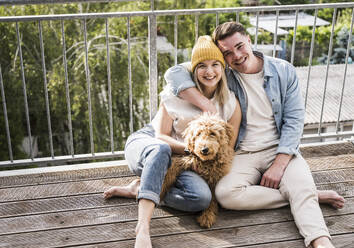Glücklicher junger Mann und Frau sitzen mit Hund auf dem Balkon - UUF28784