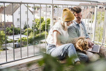 Happy couple using laptop and sitting with dog on balcony - UUF28787