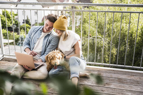 Glückliches junges Paar mit Laptop und Hund auf dem Balkon - UUF28789