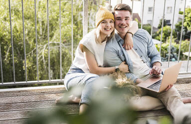 Happy couple sitting with laptop and dog on balcony - UUF28791