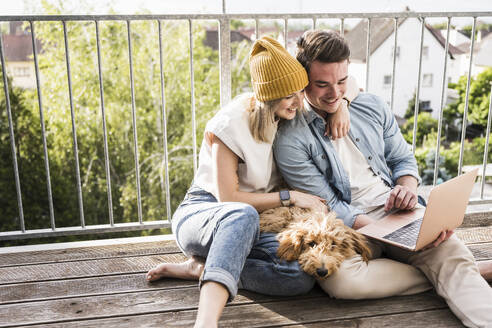 Glückliches junges Paar, das zusammen auf dem Balkon einen Laptop benutzt - UUF28792