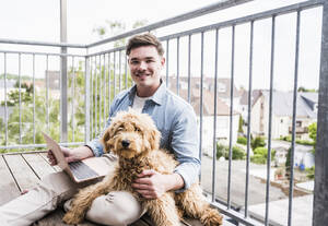Glücklicher Mann sitzt mit Hund und Laptop auf dem Balkon - UUF28814