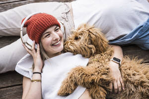 Happy woman wearing wireless headphones lying on floor - UUF28827