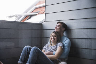Lächelnde junge Frau mit Mann an der Wand sitzend auf dem Balkon - UUF28836