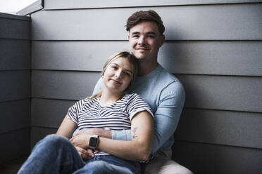 Lächelndes junges Paar sitzt zusammen an der Wand auf dem Balkon - UUF28838
