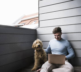 Lächelnder junger Mann mit Laptop und Hund auf dem Balkon sitzend - UUF28839